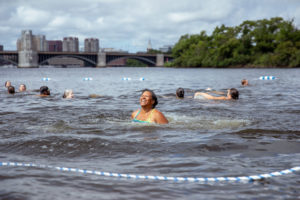 A woman smiles while swimming in a river with other swimmers nearby. There are trees, a bridge, and city buildings in the background under a cloudy sky. Blue and white ropes mark the swimming area.