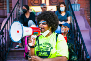 A woman wearing sunglasses and a bright yellow shirt speaks into a megaphone while sitting on outdoor steps with others, some wearing masks, during a protest or gathering.