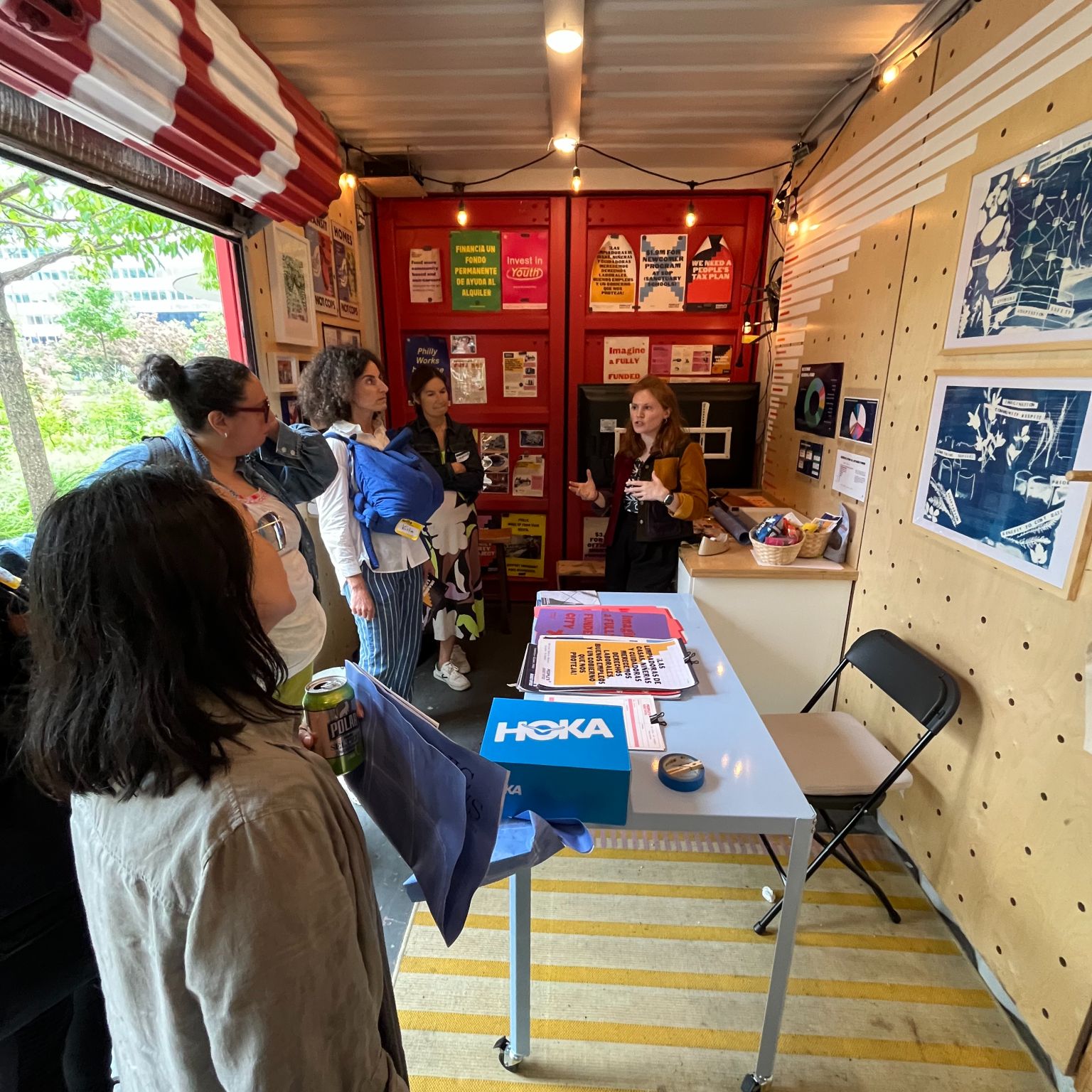 A group of people listens to a woman giving a presentation inside a small, colorful room decorated with posters and artwork on the walls. A table with papers and a shoebox is in the center of the room.