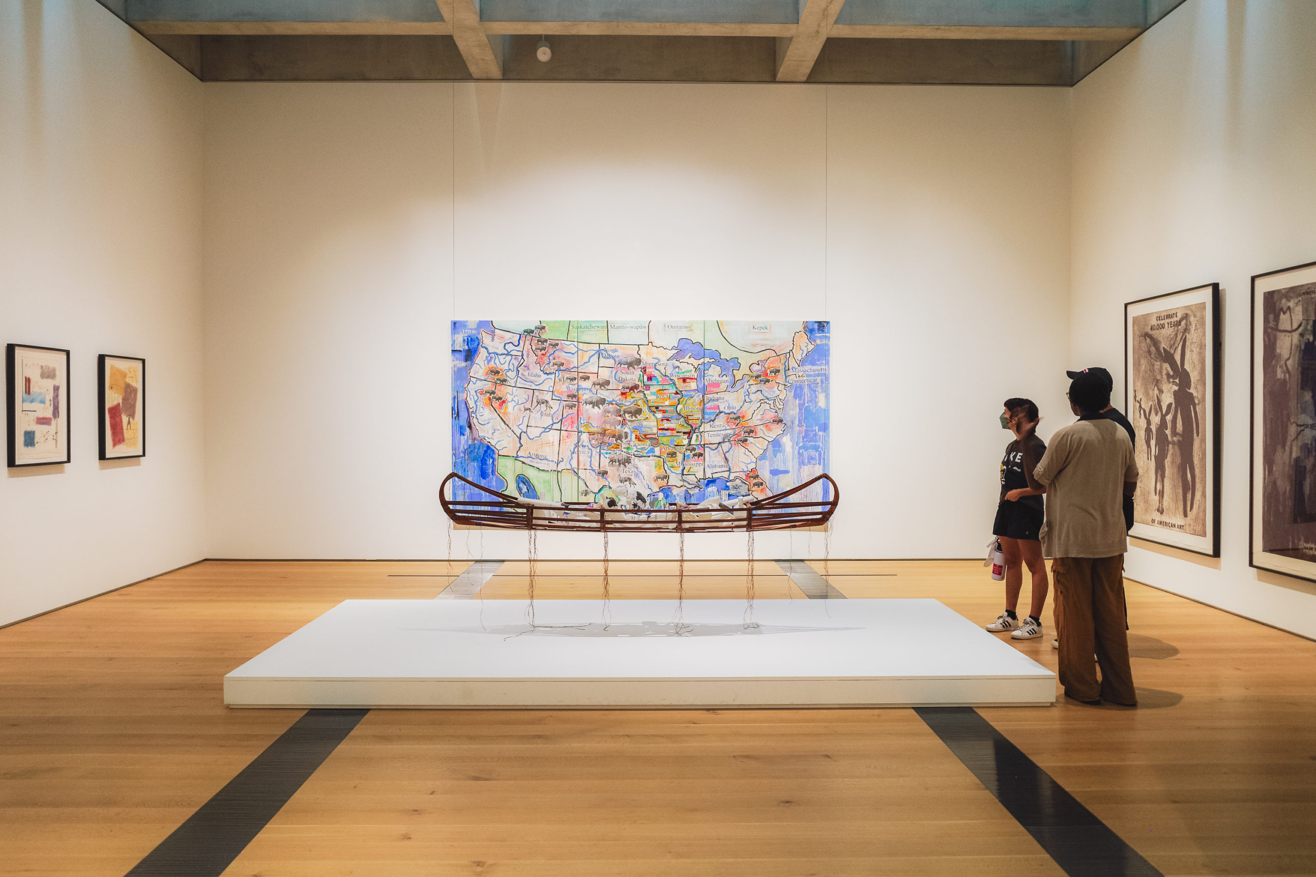 Two people view a wooden canoe sculpture on display in an art gallery, with a colorful map of the United States on the wall behind it and framed artworks on the surrounding white walls.