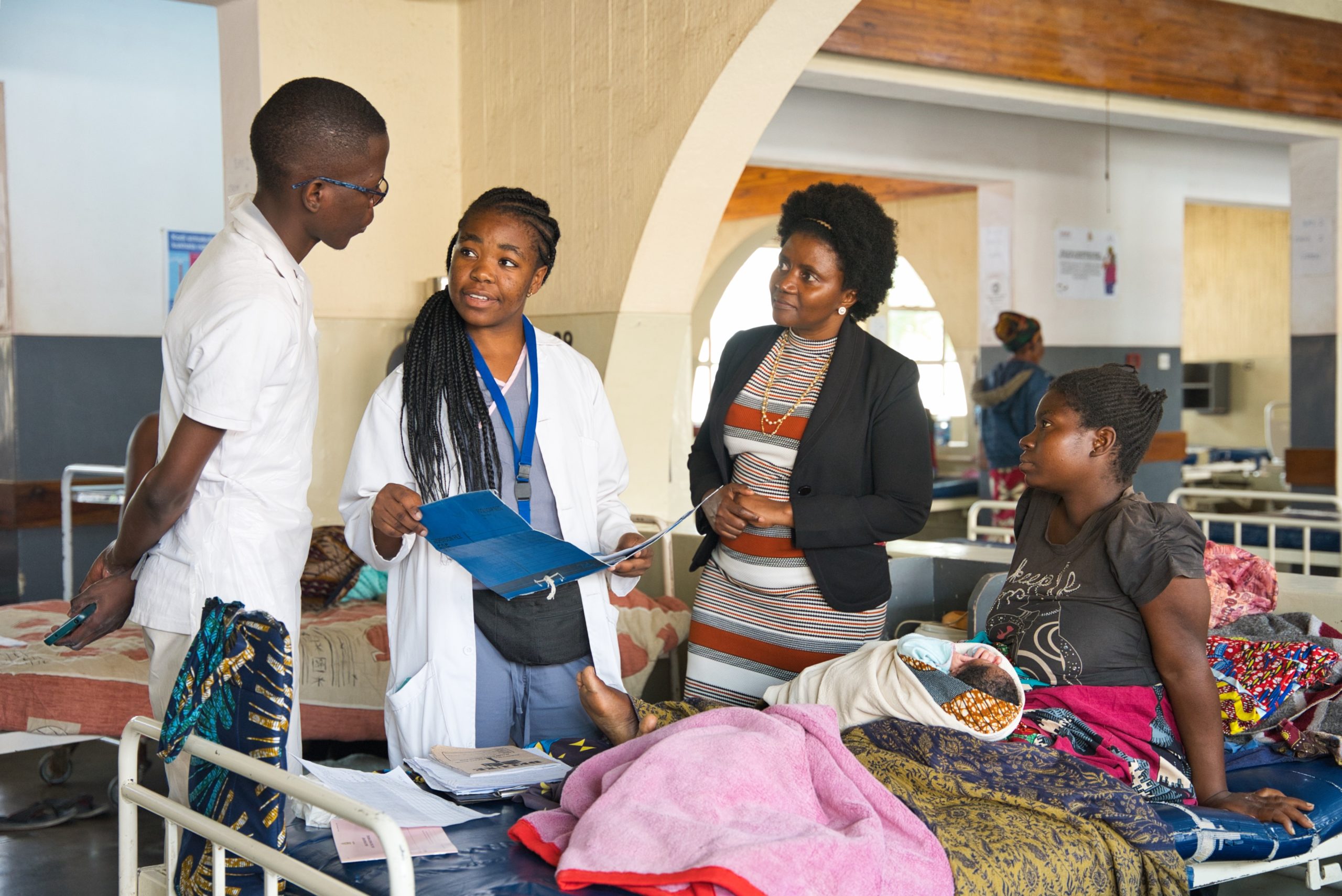 A medical team speaks with a woman and her baby in a hospital ward. Two healthcare workers, one in a white coat, hold a chart while talking to the patient and another woman, likely a visitor or social worker, standing nearby.