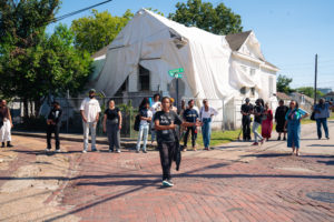 A group of people stands on a street corner near a house covered in white tarp, while one person in black walks toward the camera on a brick road under a clear blue sky.