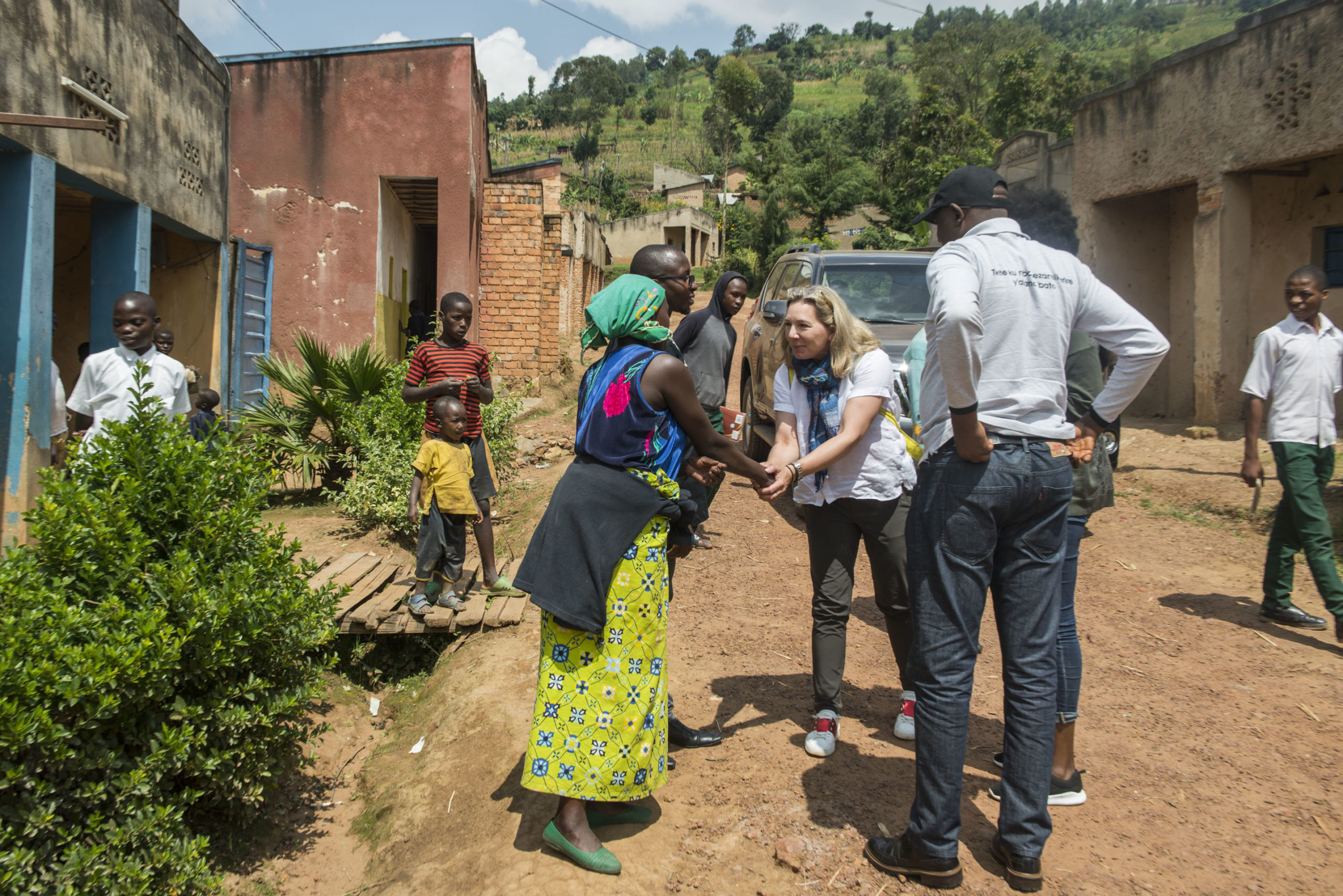 A woman in a green skirt shakes hands with a visitor in casual clothes on a dirt path, surrounded by children and other adults in a rural village setting with brick and concrete buildings.