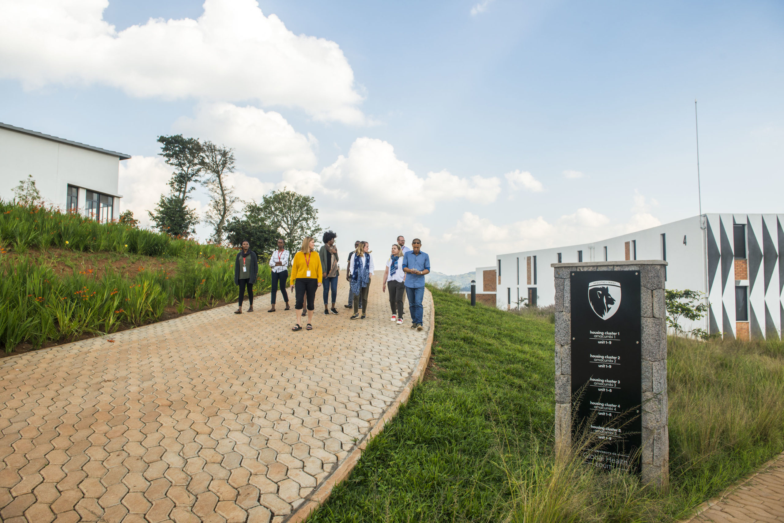A group of people walks up a paved path between modern buildings on a green campus under a partly cloudy sky, with a black signpost visible in the foreground.