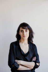 A woman with medium-length dark hair, wearing a black blouse, stands against a plain light background with her arms crossed and a neutral expression.