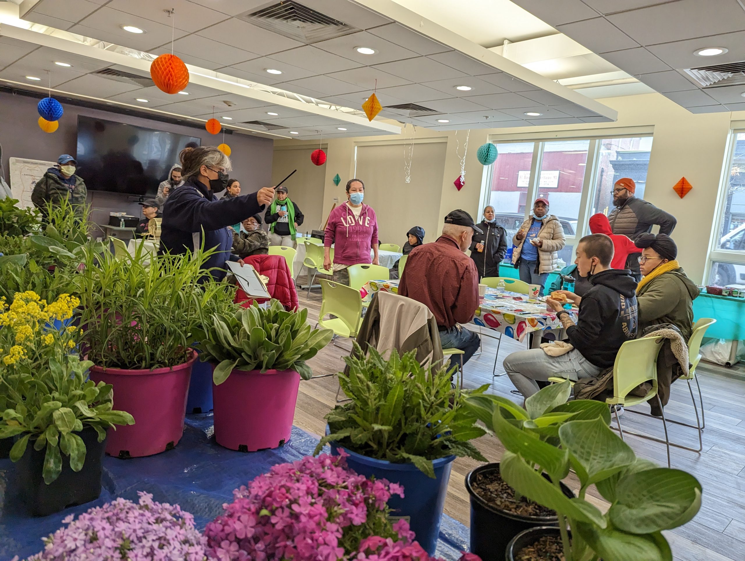 A group of people, some wearing masks, gather in a brightly lit room decorated with colorful paper lanterns. Potted flowers and plants are on tables in the foreground, while a person leads a discussion or activity.