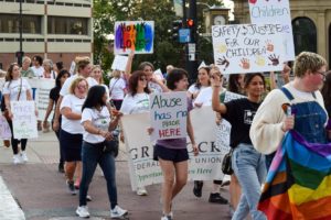 A diverse group of people march on a city street holding signs about child safety, justice, and anti-abuse. One person carries a rainbow pride flag. The atmosphere appears peaceful and supportive.
