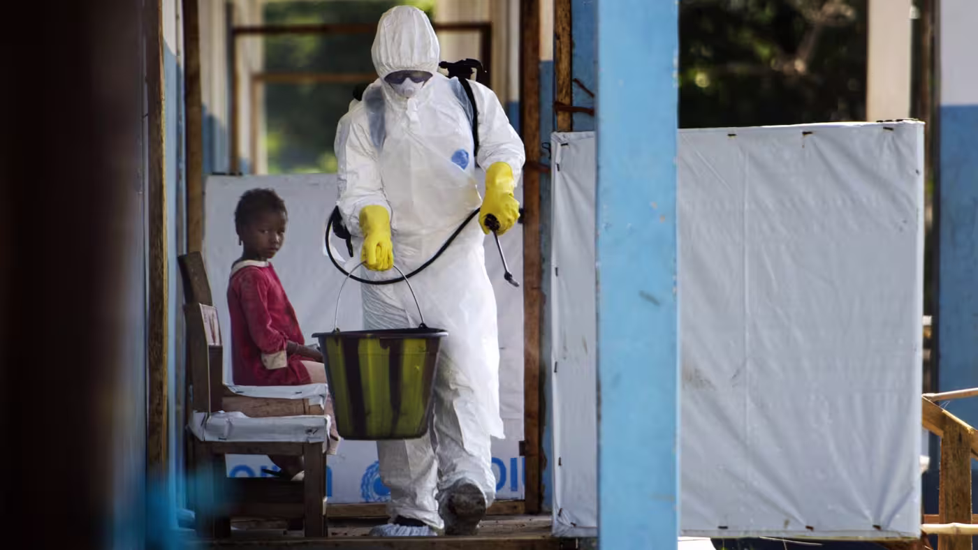 A person in full protective gear and yellow gloves carries a bucket and sprayer in a corridor, while a child in a red outfit sits on a bench nearby, watching.