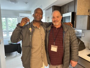 Two men stand smiling in a modern apartment. One man holds up a key with a keychain, while the other has his arm around him. There is natural light coming from a window in the background.