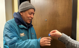 A man in a blue jacket and gray beanie smiles as he receives a set of keys from another person in front of a brown door.