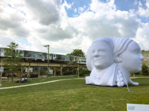 A large, white, sculptural installation featuring multiple abstract human faces sits on a grassy field near elevated train tracks, with a train passing by under a partly cloudy sky. A sign reads Floating Museum.