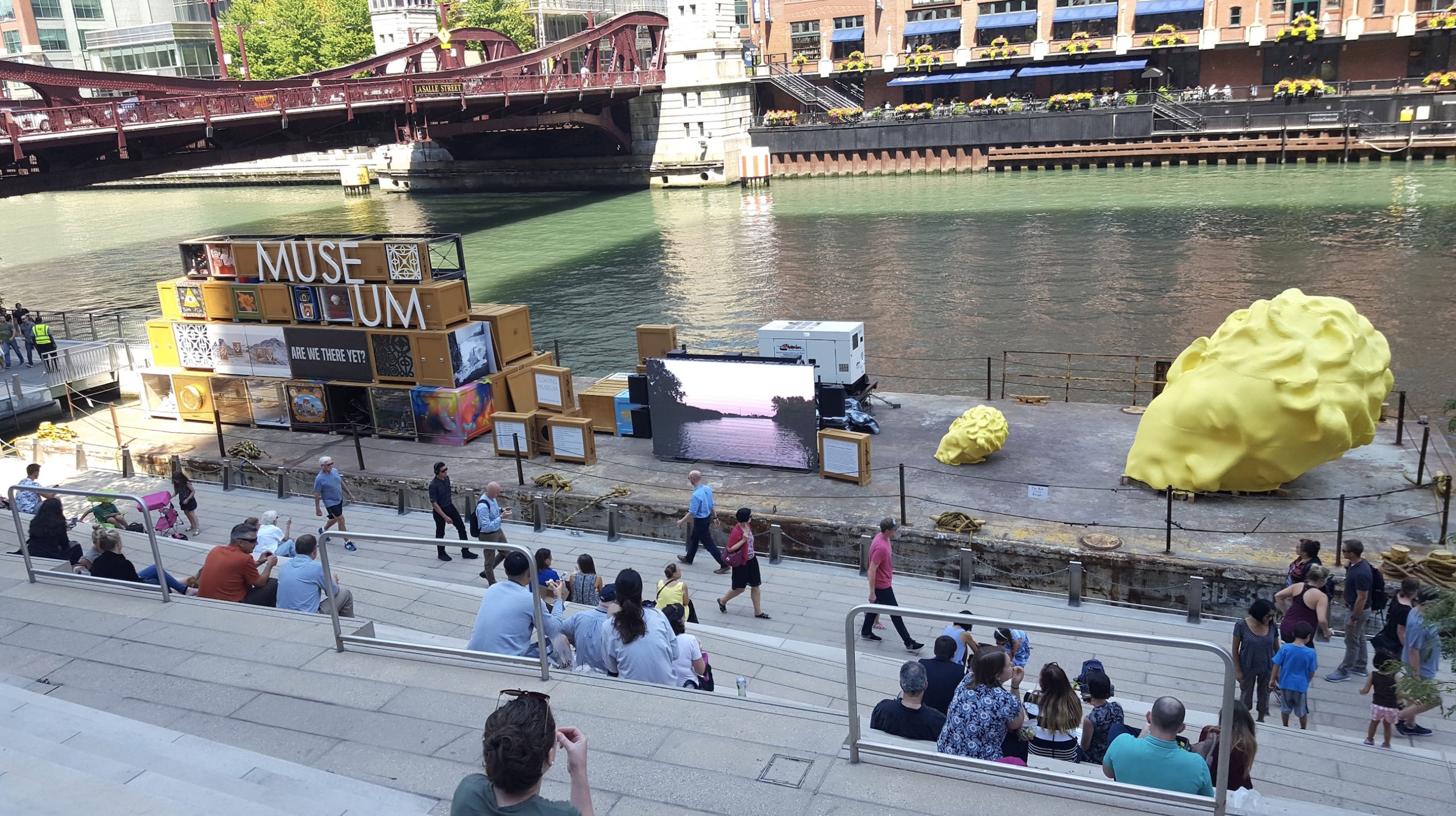 People sit on stepped seating by a riverside, facing art installations including stacked boxes labeled “MUSE UM,” digital screens, and two large yellow sculptures. A red bridge and city buildings are in the background.