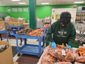 A person wearing a green Food For Free hoodie and blue gloves sorts carrots in a food distribution center with bins of vegetables and boxes around them. A Food For Free sign is on the wall in the background.