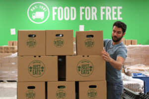 A man stacks cardboard boxes labeled Just Eats in front of a green wall with the words Food For Free and a truck graphic. Pallets and packaging materials are visible in the background.