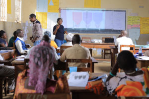 A group of adults sit at desks in a classroom, listening to two presenters at the front. A projector displays information on a screen, and colorful posters and notes decorate the walls.