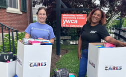 Two women stand outdoors by boxes labeled CARES, filled with toiletries and supplies, in front of a YWCA sign that reads eliminating racism, empowering women. They are smiling.
