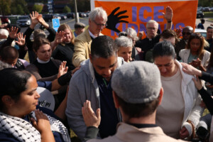 A group of people stand close together with eyes closed and hands raised in prayer or support. In the background, an orange sign with the words ICE OUT and a black hand is visible.