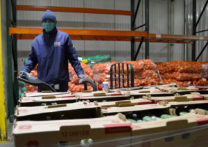 A person wearing a blue jacket, hat, and face mask operates a pallet jack in a warehouse filled with boxes of produce and large sacks of onions.