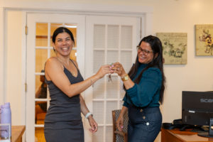 Two women smiling and holding a set of keys together in a home office setting. One woman wears a dark dress; the other wears a blue blouse and glasses. Paintings and a computer are visible in the background.