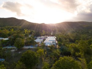 Aerial view of a small village surrounded by lush green trees and hills at sunrise or sunset, with sunlight streaming through the clouds in the background.