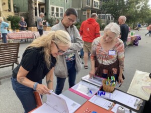 Three women stand at an outdoor table covered with papers, binders, and office supplies at a neighborhood event; one woman points at a binder while the others look on attentively. People and houses are visible in the background.