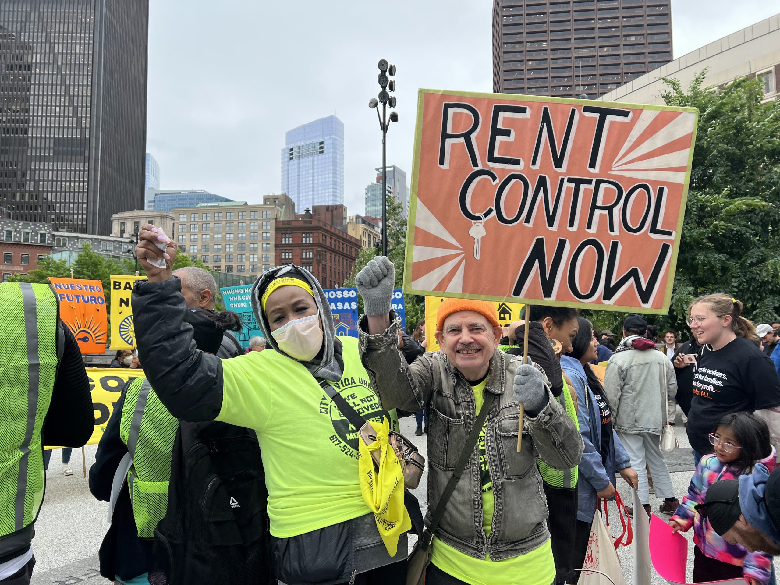 Two people at a rally hold a large sign reading “RENT CONTROL NOW.” Both wear bright yellow shirts and vests, and one wears a face mask. Other protesters and city buildings are visible in the background.