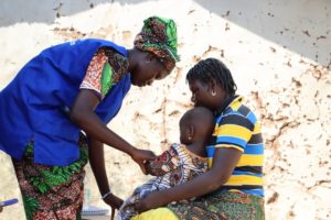 A healthcare worker in a blue vest administers a vaccine to a young child sitting on a woman’s lap. The woman comforts the child, who is wearing patterned clothing. They are outdoors against a light-worn wall.