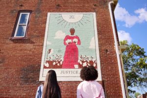 Two people with long hair stand facing a mural painted on a brick wall. The mural depicts a woman in a red dress holding two white doves, surrounded by cotton plants, with JUSTICE written at the bottom.