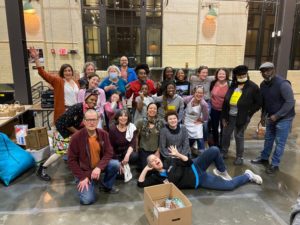 A diverse group of people pose and smile together indoors, some standing and some kneeling on the floor. There are food items and boxes nearby, suggesting a community event or volunteer gathering.