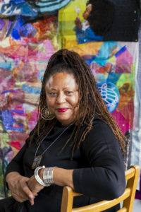 A woman with long dreadlocks, wearing a black outfit, silver jewelry, and large earrings, sits confidently on a wooden chair. A colorful, abstract textile artwork serves as the vibrant background.