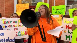 A young person in an orange jacket speaks into a megaphone while holding papers, surrounded by others holding colorful protest signs in Spanish in front of a brick building.