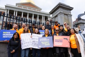 A diverse group of people hold signs about ending hunger, including “Make Hunger History” and “Every Child Deserves Dinner,” standing in front of a government building with columns and a gold dome.