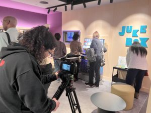 A person films with a video camera on a tripod while several people interact with a digital jukebox display on a wall labeled JUKE BOX in a modern, well-lit room.