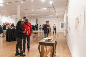 People observe artwork in a bright, modern gallery. A man and woman stand together looking at art on the wall, while another man examines a piece on a table. Several other visitors are scattered throughout the room.