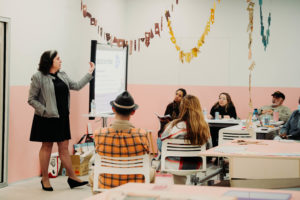 A woman in business attire stands and points at a presentation screen while a group of seated adults listens attentively in a classroom decorated with colorful hanging banners.