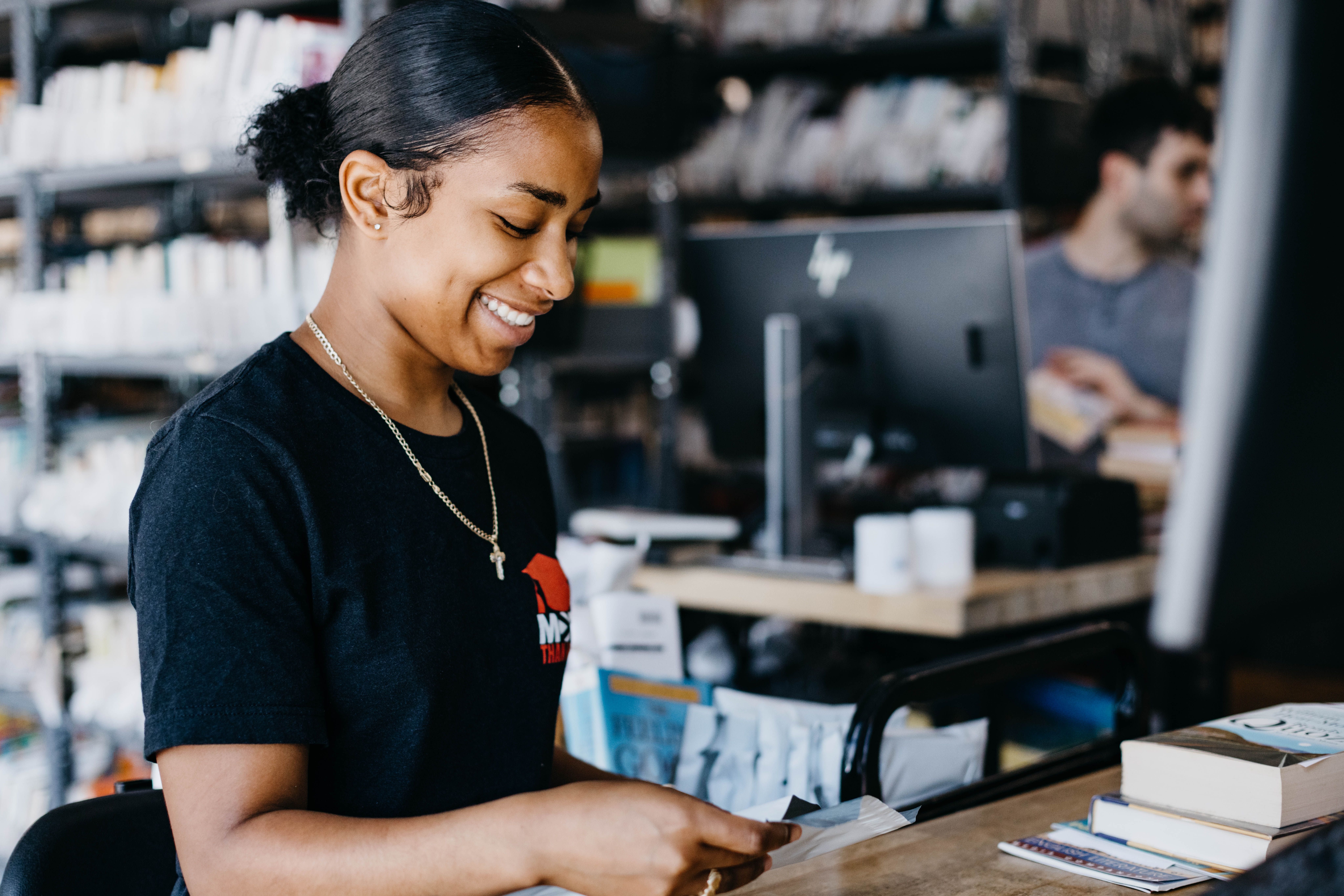 A smiling woman wearing a black t-shirt stands behind a counter in a store, handling papers. Shelves with various products and another person at a computer are visible in the background.