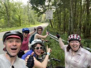 Four smiling cyclists wearing helmets pose and point at a road sign that reads Motorists Give 4 Ft to Pass on a rural roadside surrounded by trees.