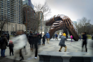 People walk around and take photos of a large bronze sculpture of two arms embracing, situated in a city park surrounded by tall buildings and leafless trees on a cloudy day.