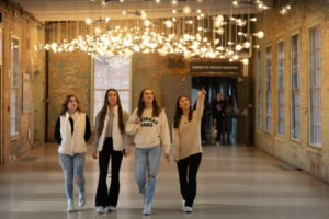 Four young women walk together in a hallway with exposed brick walls and many hanging lightbulbs above them; one woman is pointing upward, and others look in various directions.