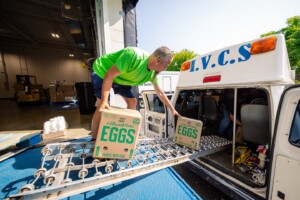 A man in a green shirt loads cartons labeled EGGS into the back of a white van at a warehouse loading dock, using a conveyor ramp. The van has V.C.S written on it in blue letters.