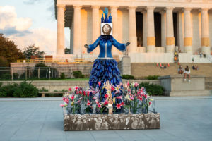 A colorful statue of a joyful woman in a blue dress stands in front of the Lincoln Memorial, surrounded by pink flowers and black-and-white images of people at its base.