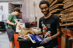 A young person wearing gloves and a black t-shirt smiles while holding a blue book in a storage area with cardboard boxes. Another person in the background sorts items near open containers.