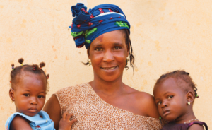 A woman in patterned clothing and a blue headscarf smiles, standing in front of a yellow wall, with two young girls in blue dresses, each standing on either side and gently holding onto her.