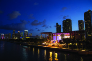 A vibrant cityscape at dusk with illuminated skyscrapers, a modern waterfront building with colorful lights, and reflections on the calm water under a deep blue sky.