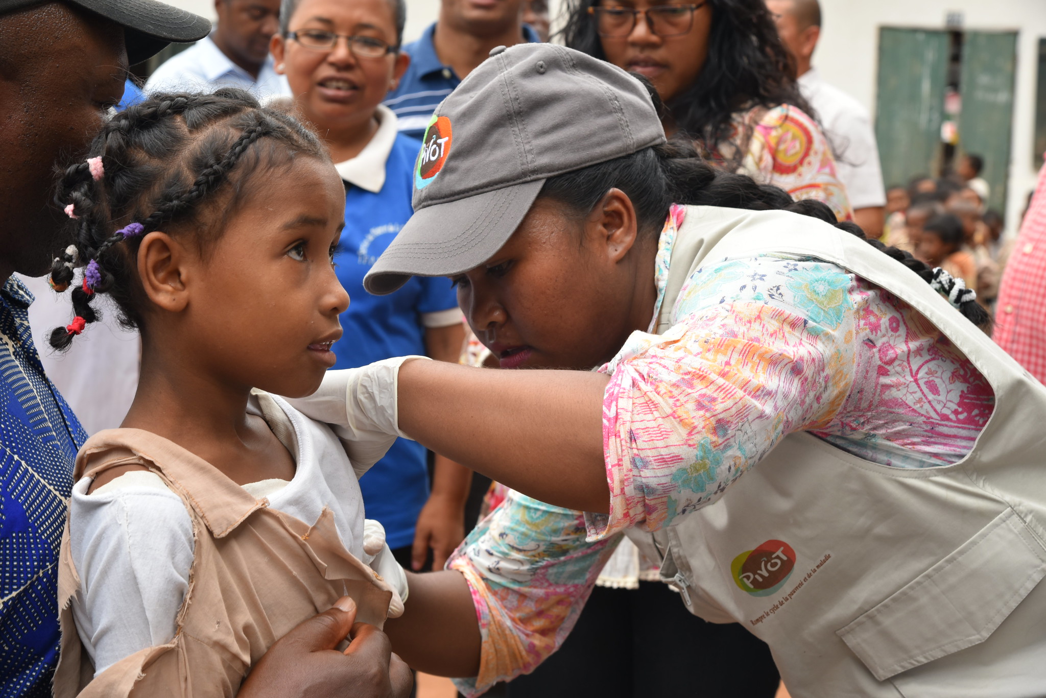A healthcare worker in a cap and gloves gives a vaccination to a young girl with braided hair, surrounded by other adults and children in an outdoor setting.