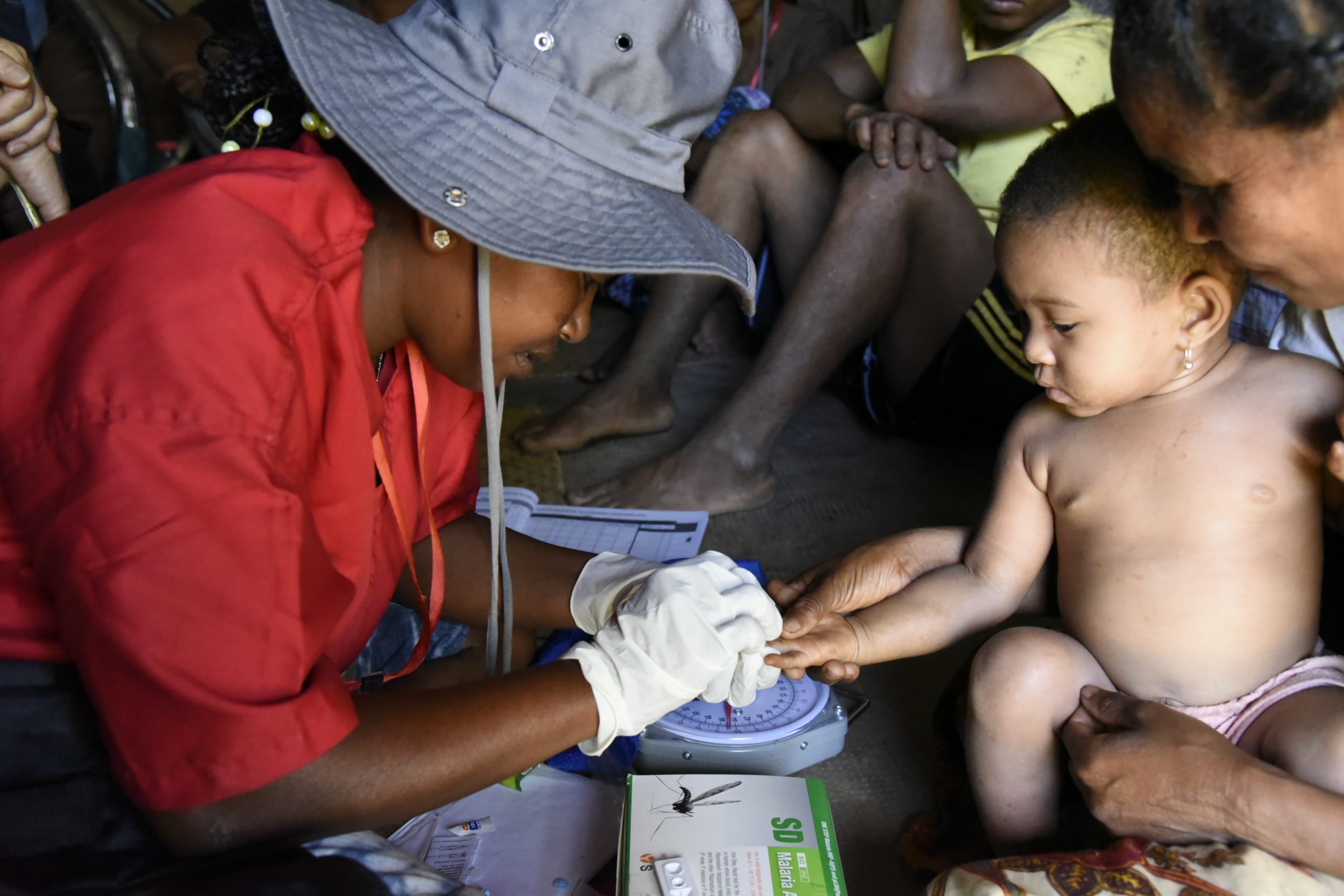 A healthcare worker in a red shirt and gray hat pricks a young childs finger for a blood test as the child sits on an adults lap. Others are seated nearby, observing the process in a community setting.