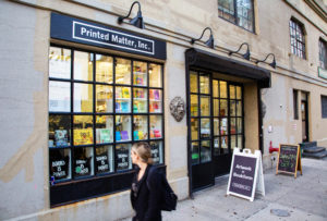 A person walks past the storefront of Printed Matter, Inc., a bookstore with large windows displaying colorful books and signs, including Books is Power, on a city sidewalk.