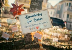 A wooden sign reading Remembering Our Babies - Propa City Community Outreach stands among white roses and small cards in a garden, honoring lost babies. Sunlight filters through autumn leaves in the background.