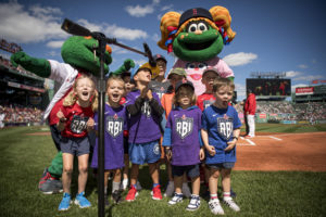 A group of smiling children in sports uniforms stand on a baseball field, with two green mascots behind them. The kids are gathered around a microphone, and a stadium crowd is visible in the background on a sunny day.
