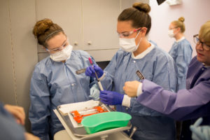 Four people in protective gowns, masks, and gloves work together in a laboratory. Two are handling pipettes and lab equipment on a tray, while the others observe and assist.
